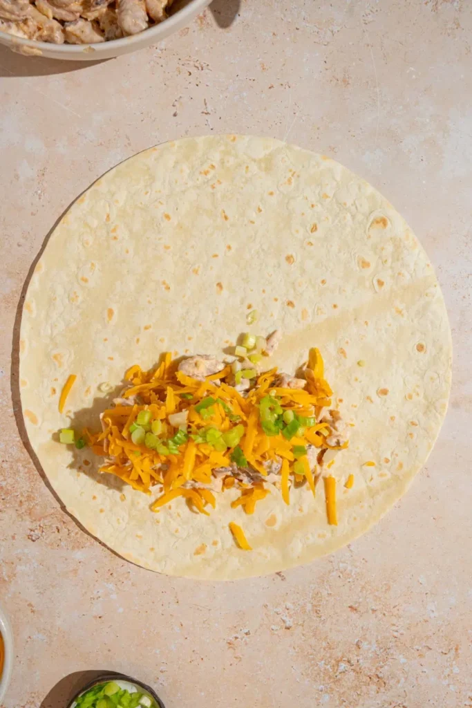 Lay one tortilla flat on a clean surface. Sprinkle about 2 tablespoons of grated cheese along the lower third of the tortilla. Spoon about ⅔ cup of the chicken mixture on top. Add another tablespoon of cheese and a sprinkle of chopped green onion.